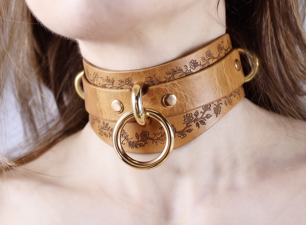 A close-up image of a woman wearing a brown leather choker necklace with a gold ring and floral design.