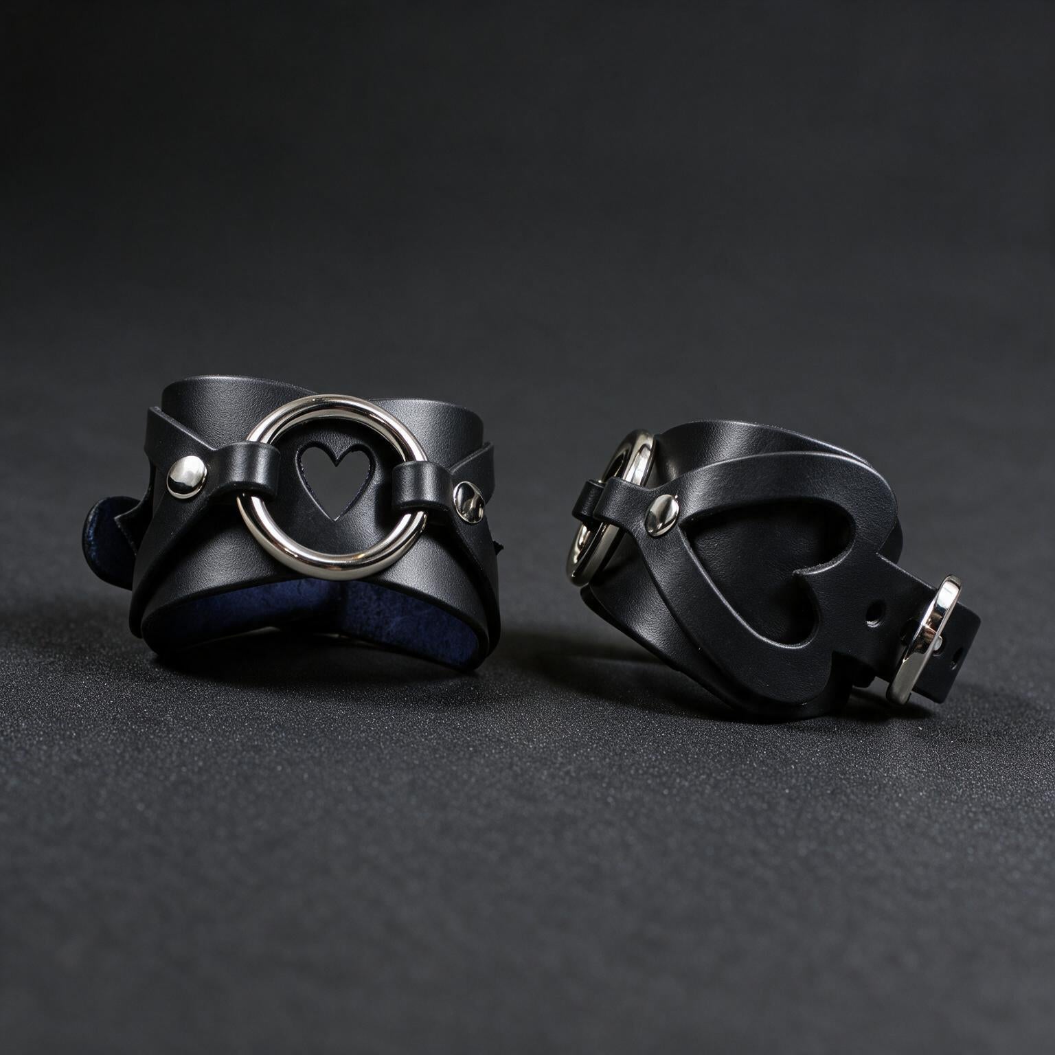 two black leather cuffs with heart-shaped holes, placed on a dark surface.