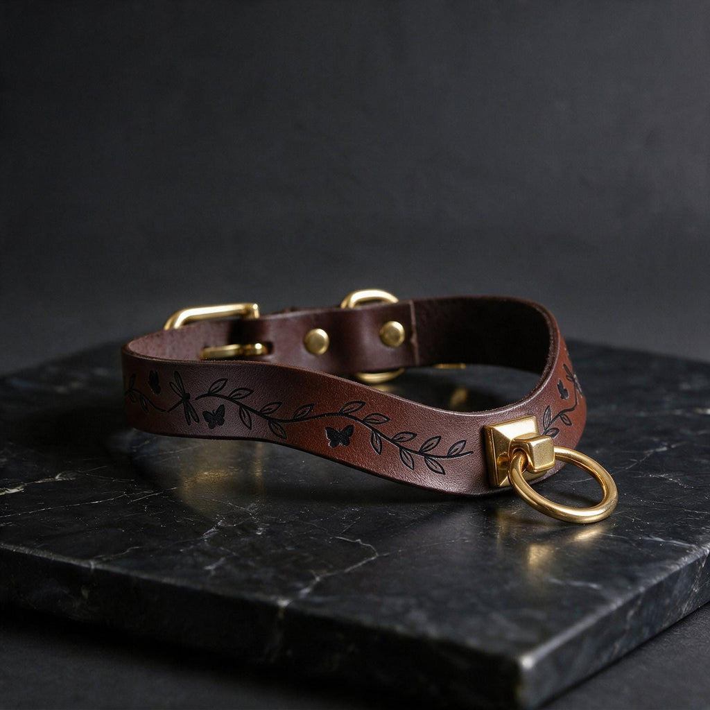 A brown leather collar with a leaf design and a gold-colored metal ring is displayed on a black marble surface.