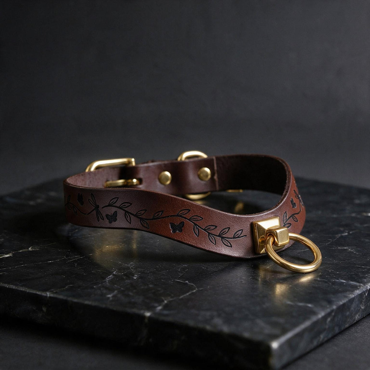 A brown leather collar with a leaf design and a gold-colored metal ring is displayed on a black marble surface.