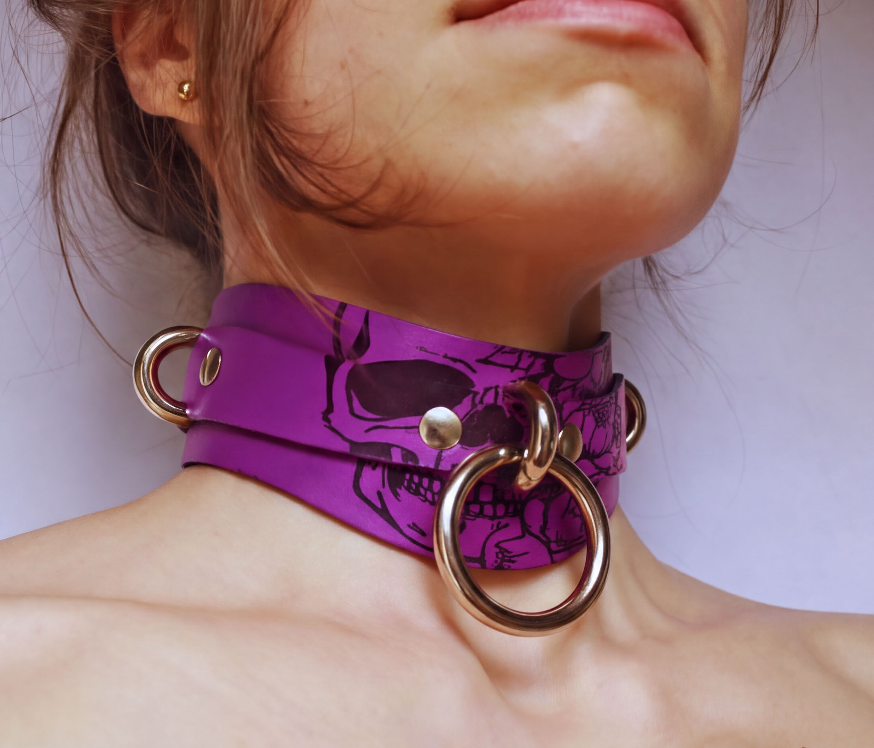 A close-up of a person's neck and chest area wearing a purple choker necklace with a skull design and metal rings.