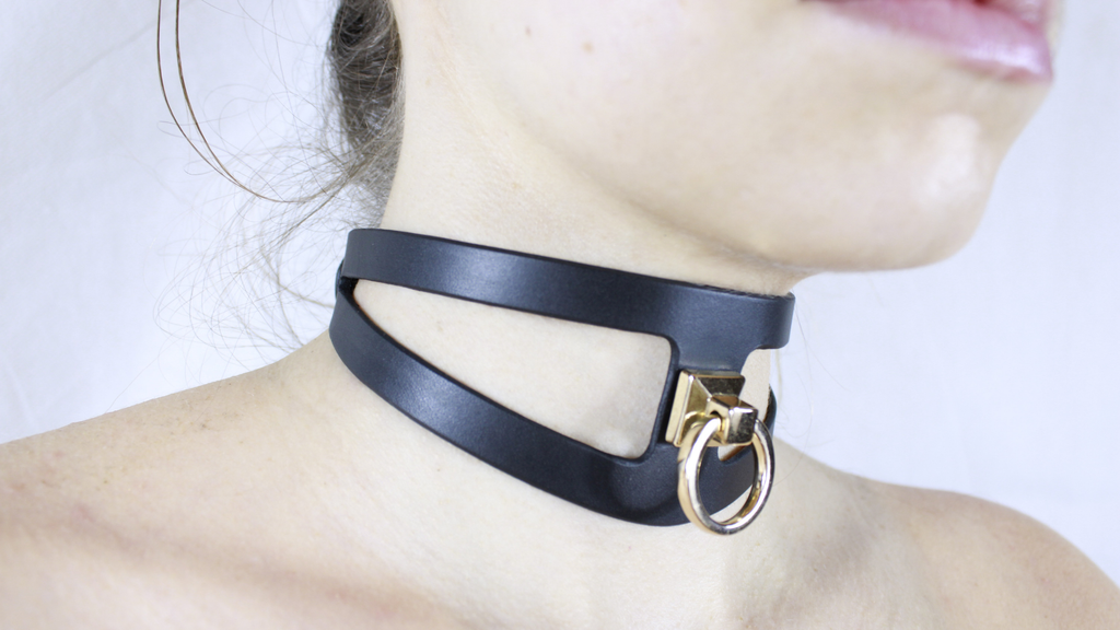 A close-up of a person's neck wearing a black choker necklace with a gold-colored metal ring.