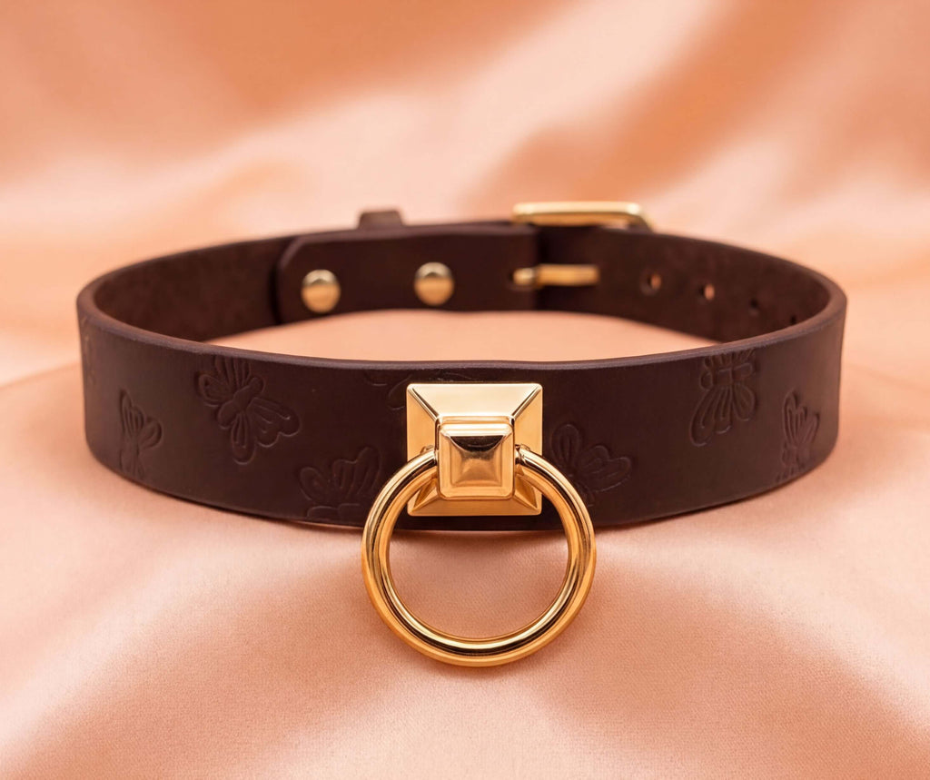 A brown leather choker collar with a gold-colored metal ring and a small square cutout on the front.