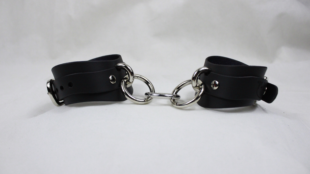 A pair of black leather cuffs with silver metal rings and buckles, arranged in a straight line on a white background.