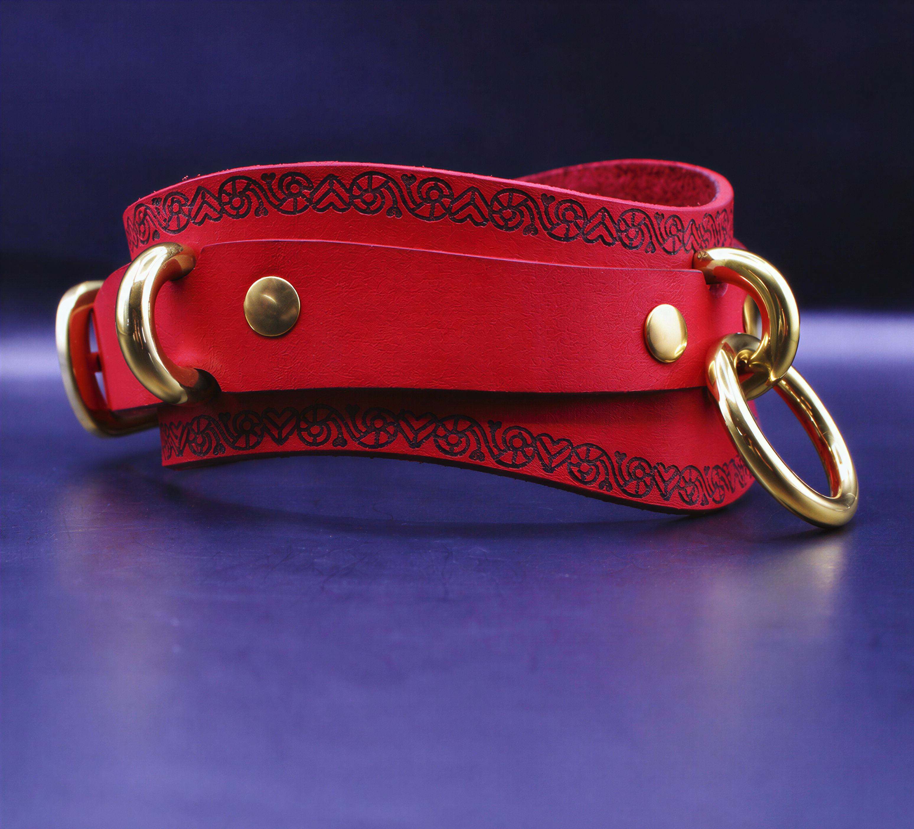 A red leather choker with gold metal rings and a decorative pattern on the edges is displayed against a dark blue background.