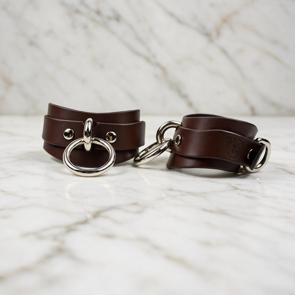 Two brown leather straps with metal rings and buckles are displayed on a marble surface.