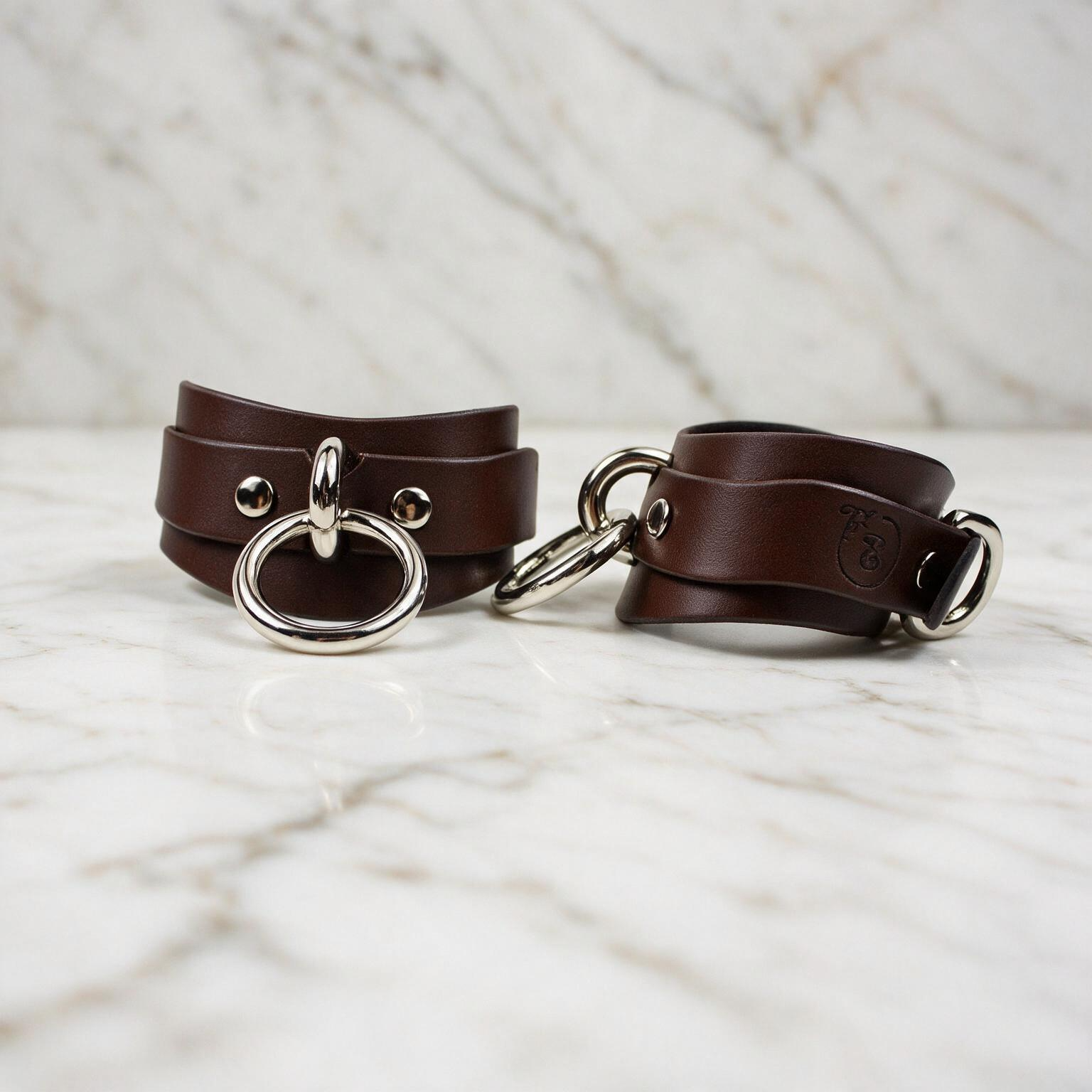Two brown leather straps with metal rings and buckles are displayed on a marble surface.