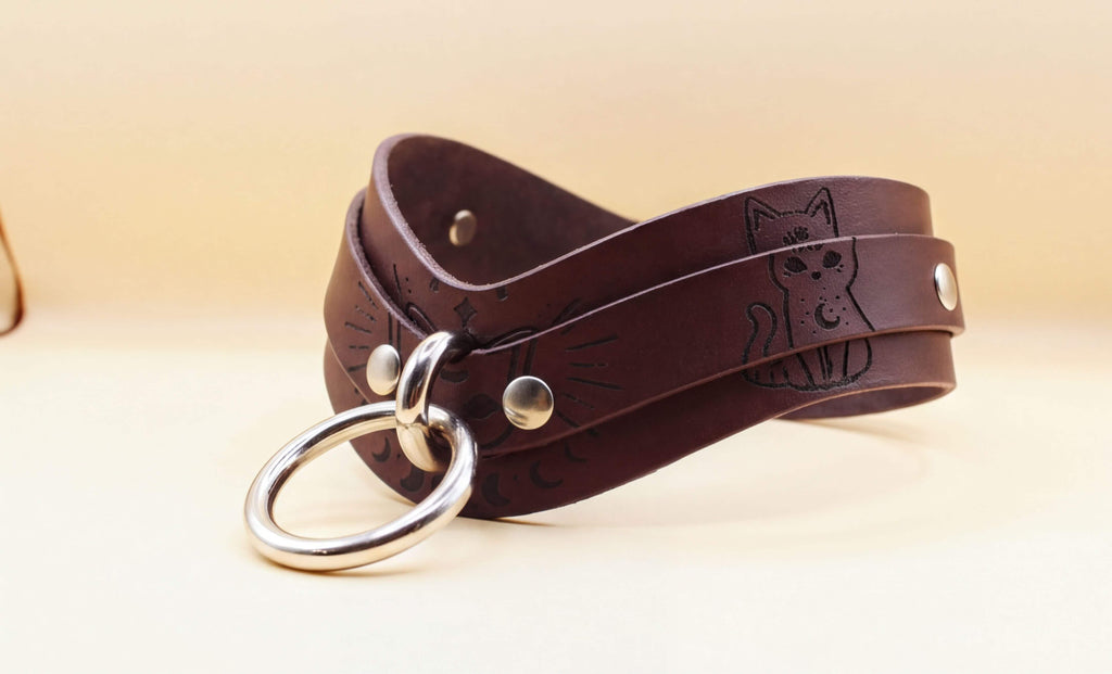 A brown leather choker necklace with a metal ring and a cat design on it.