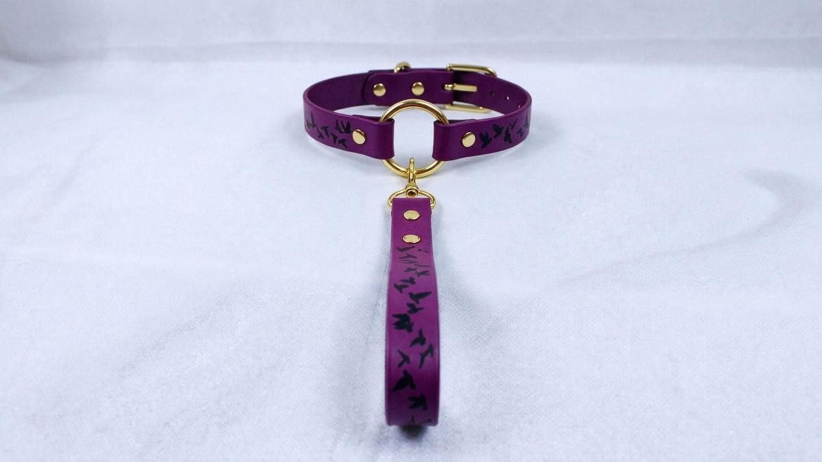 A purple collar with a gold buckle and a black bird design on it, hanging from a gold loop.