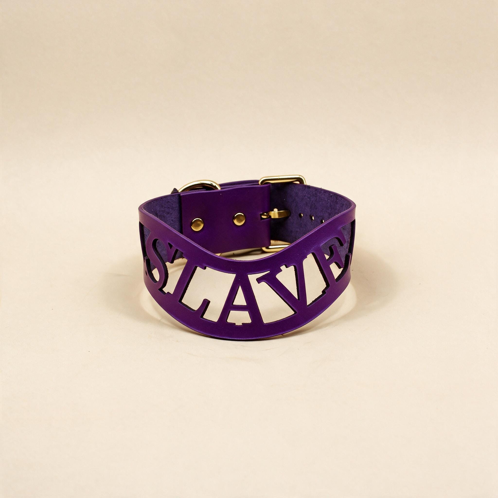 A purple leather choker necklace with the word "SLAVE" printed on it in a bold, sans-serif font.