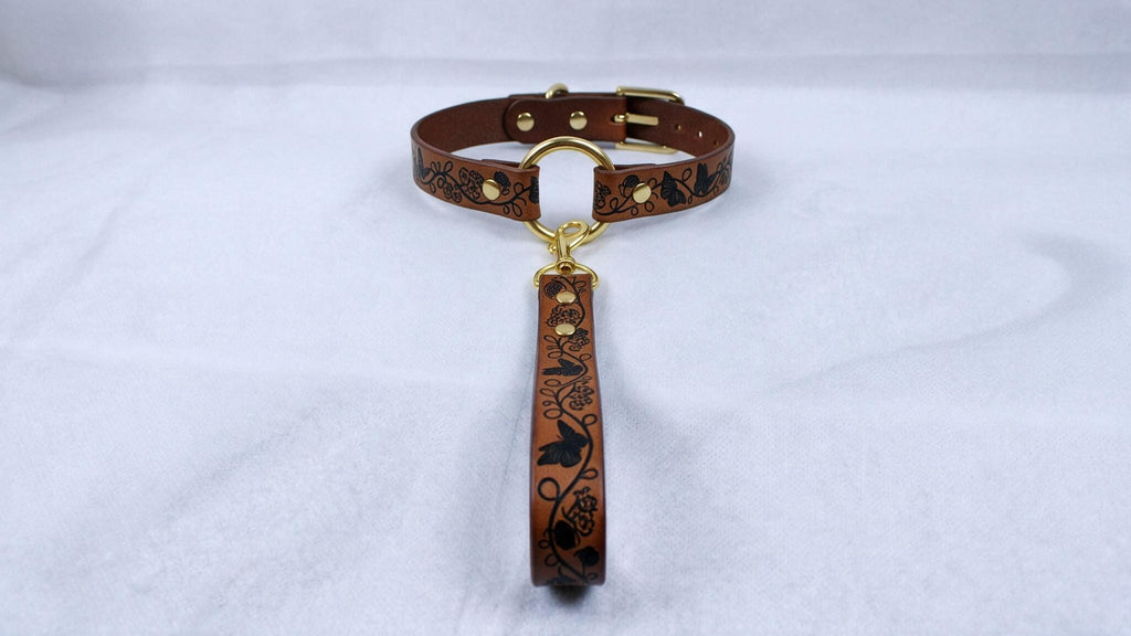 A brown leather collar with a gold buckle and a black floral pattern on the strap.