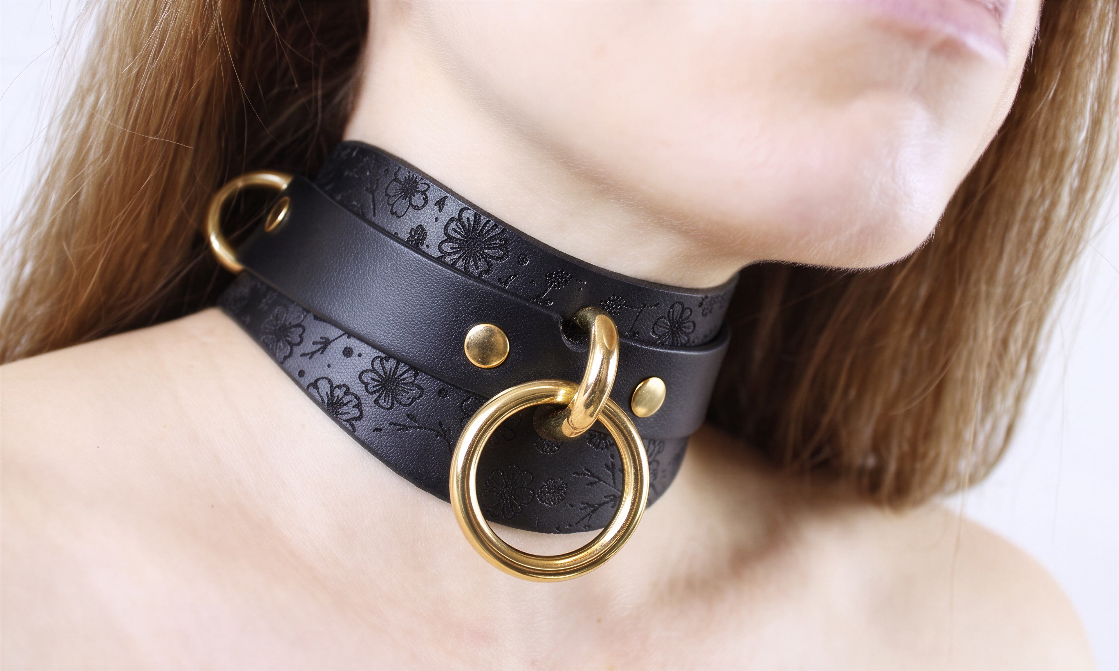 A close-up of a person's neck wearing a black leather choker necklace with gold rings and a floral pattern.
