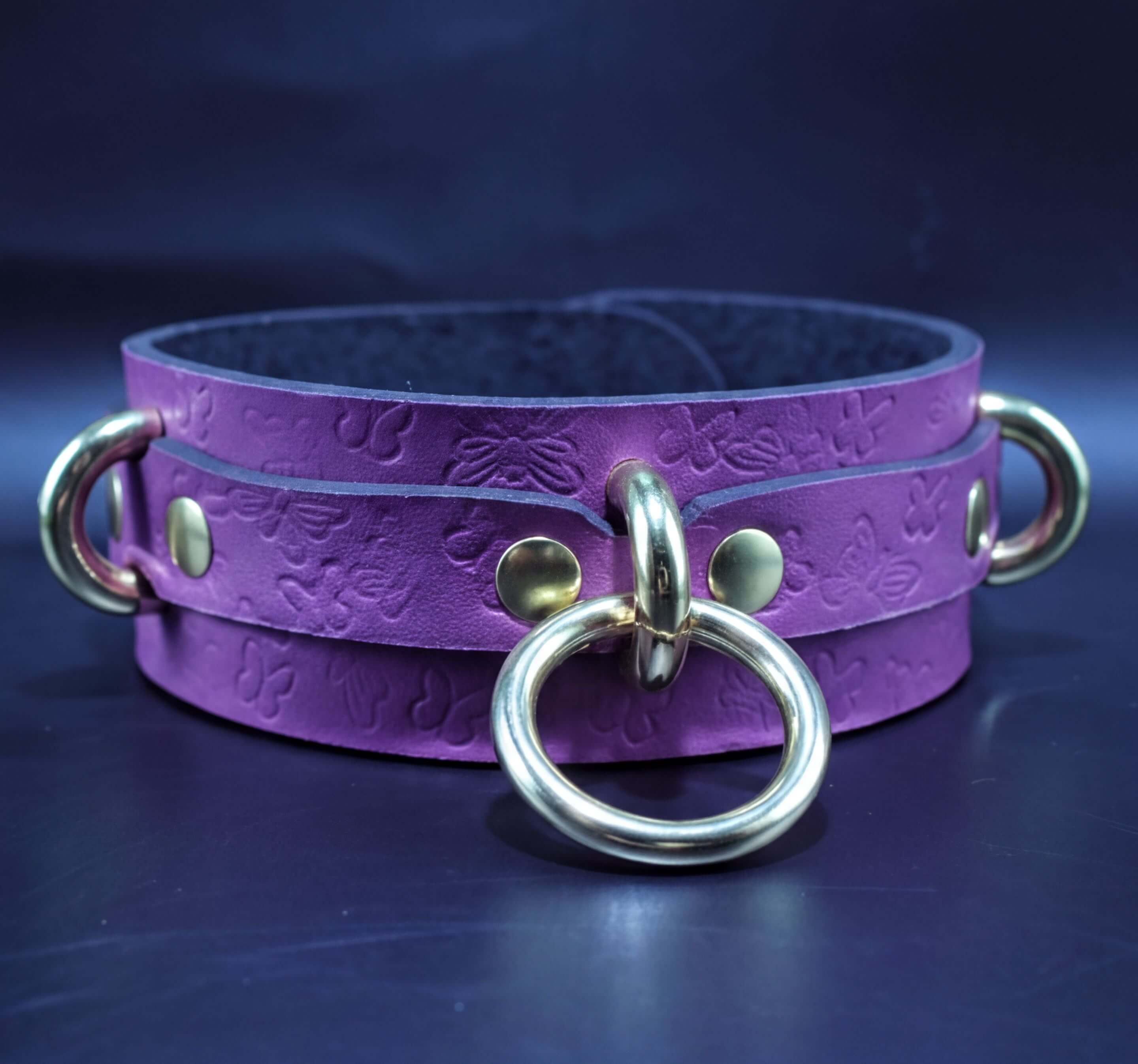 A purple leather choker collar with gold metal rings and a silver ring on the front, displayed against a dark background.
