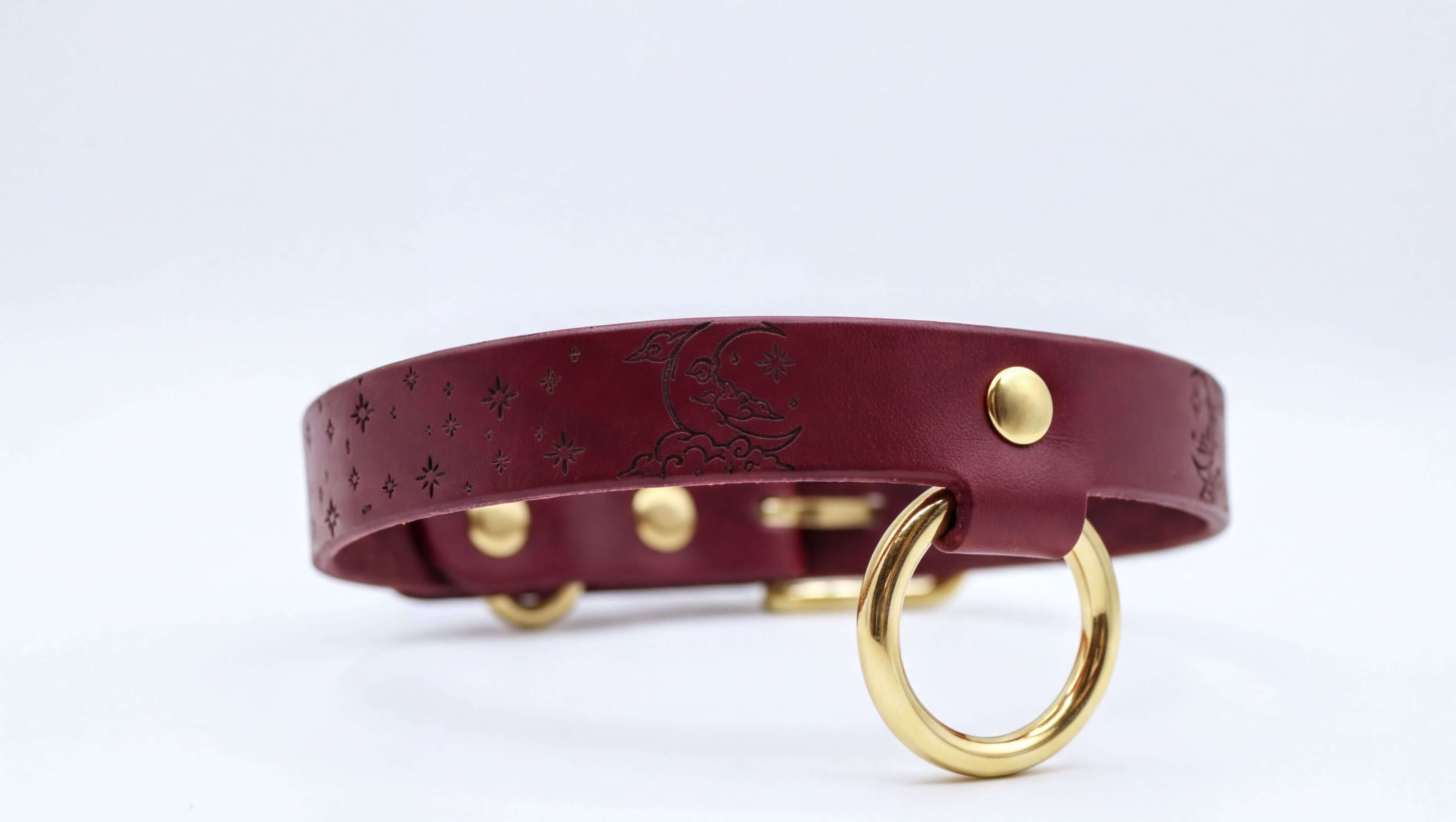 A burgundy leather collar with a starry night sky design and a gold-colored metal ring.