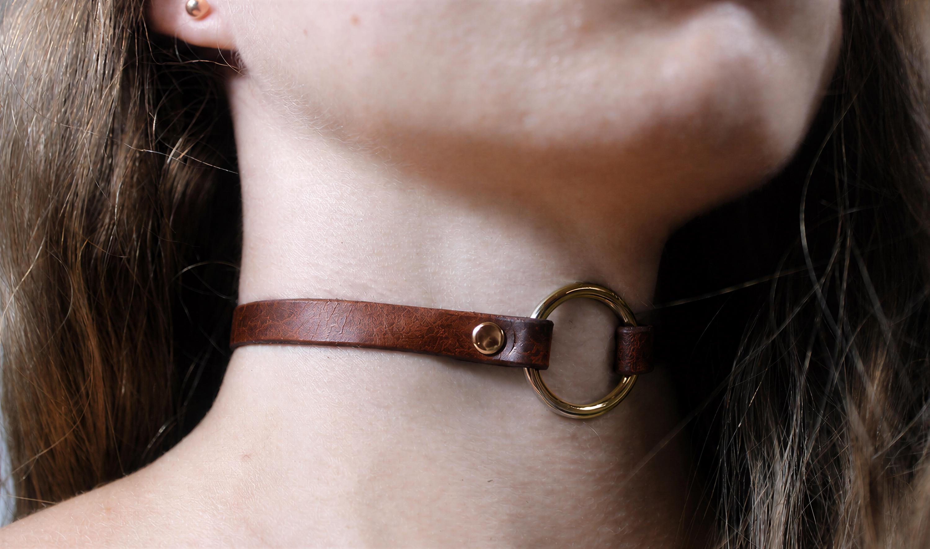 A close-up view of a woman's neck and chin area, featuring a brown leather choker necklace with a gold-colored circular clasp.