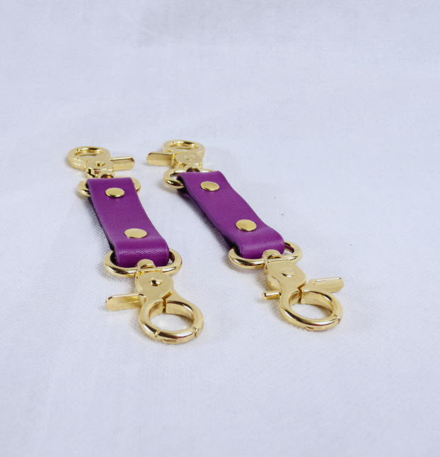 A pair of purple leather straps with gold metal clasps and buckles, placed on a white surface.