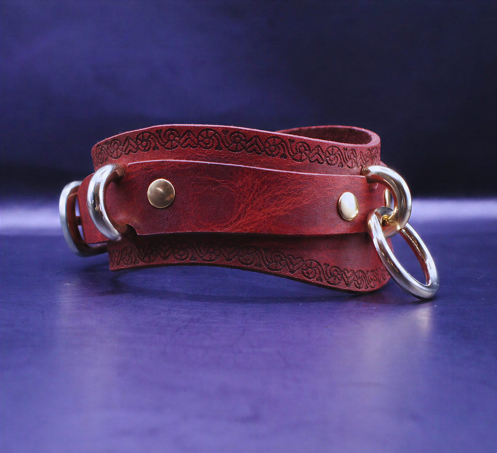 A red leather choker necklace with metal rings and a decorative pattern on the clasp is displayed against a dark blue background.