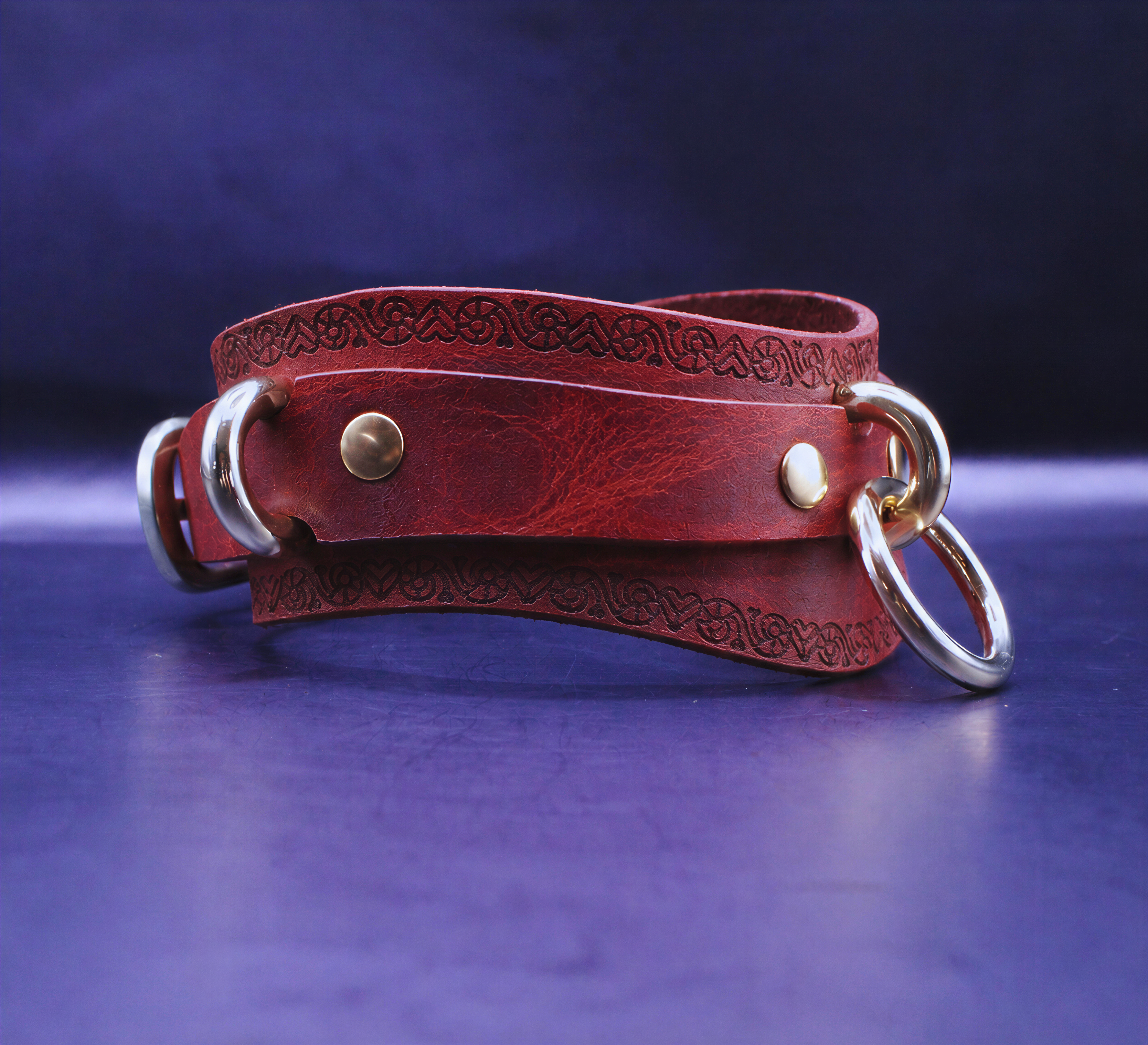 A red leather choker necklace with metal rings and a decorative pattern on the clasp is displayed against a dark blue background.