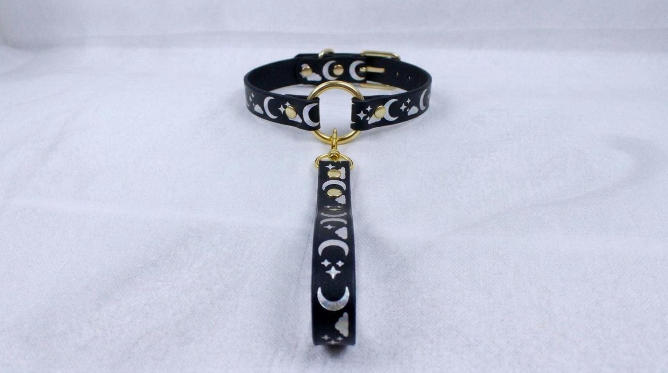 A black collar with a gold-colored metal clasp and a dangling charm featuring a moon and star design.