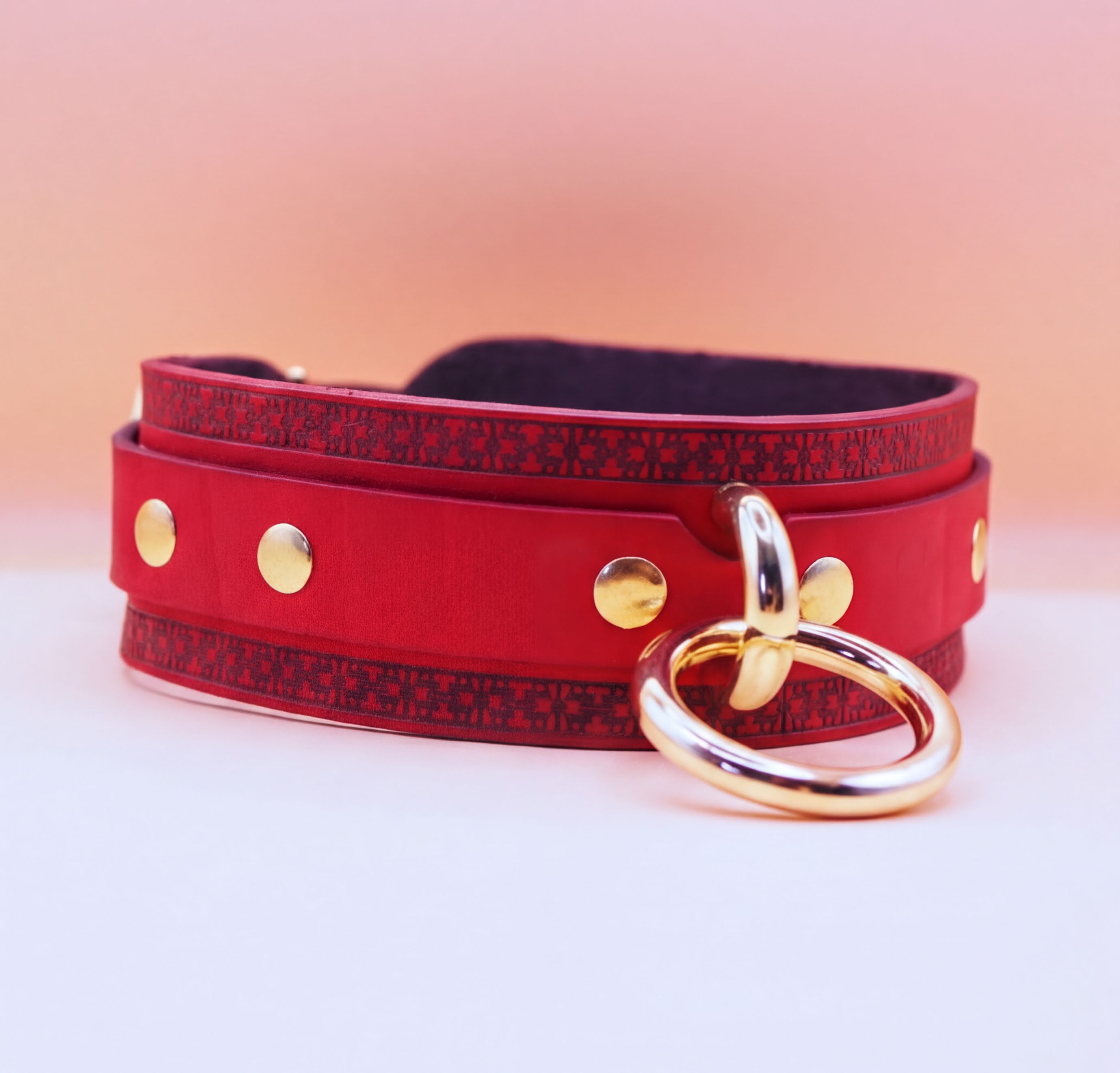 A red leather choker necklace with a gold ring and a gold buckle is displayed against a pink background.