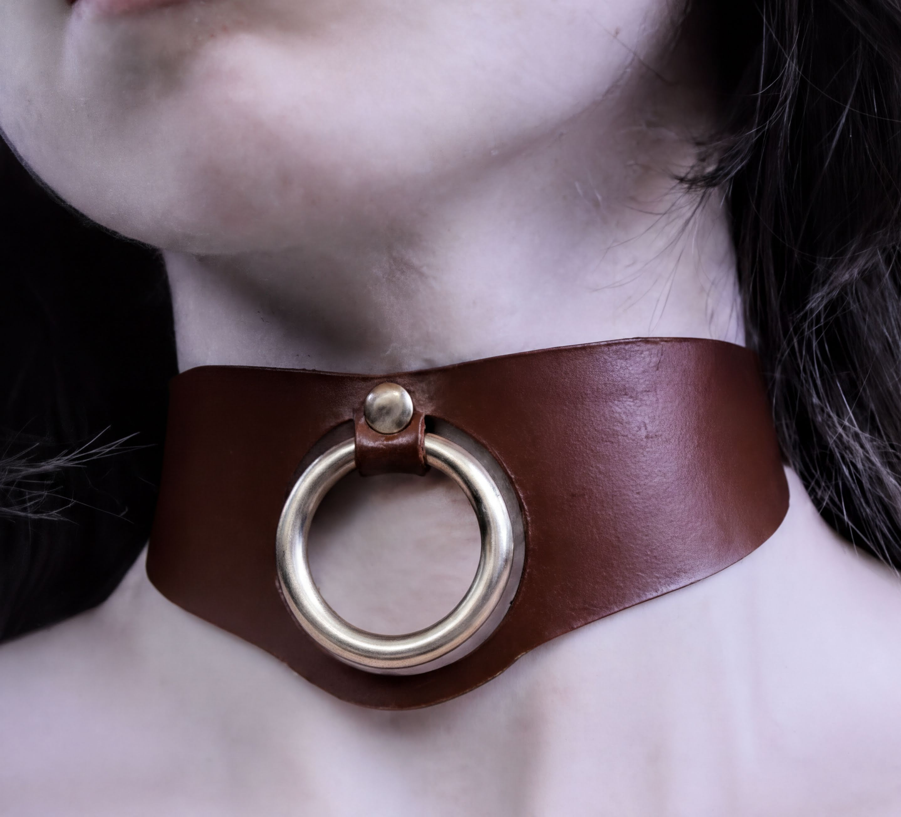 A close-up of a person's neck wearing a brown leather choker necklace with a gold-colored ring on the front.
