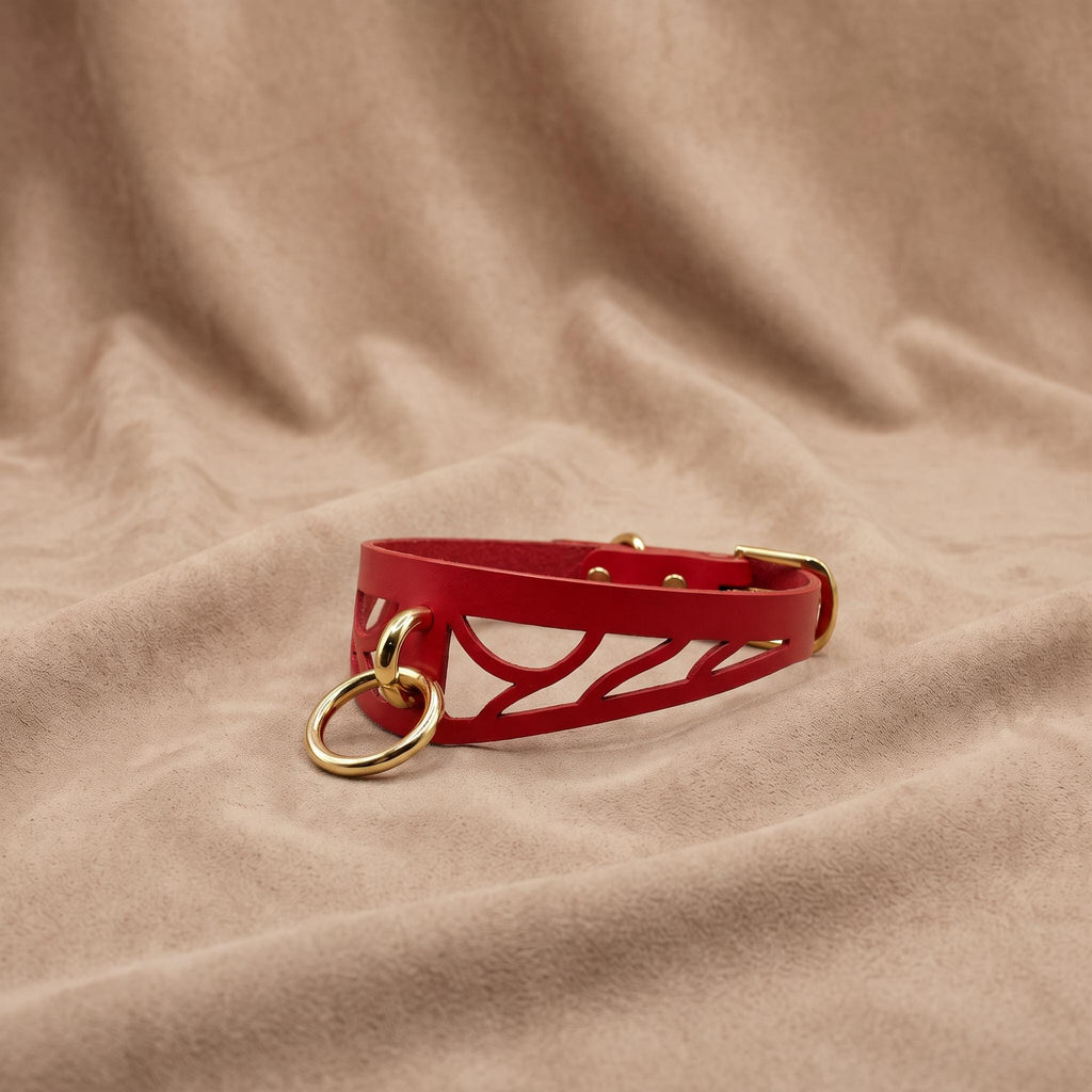 A red leather choker necklace with a cutout design and a gold-colored metal ring is displayed on a beige fabric background.