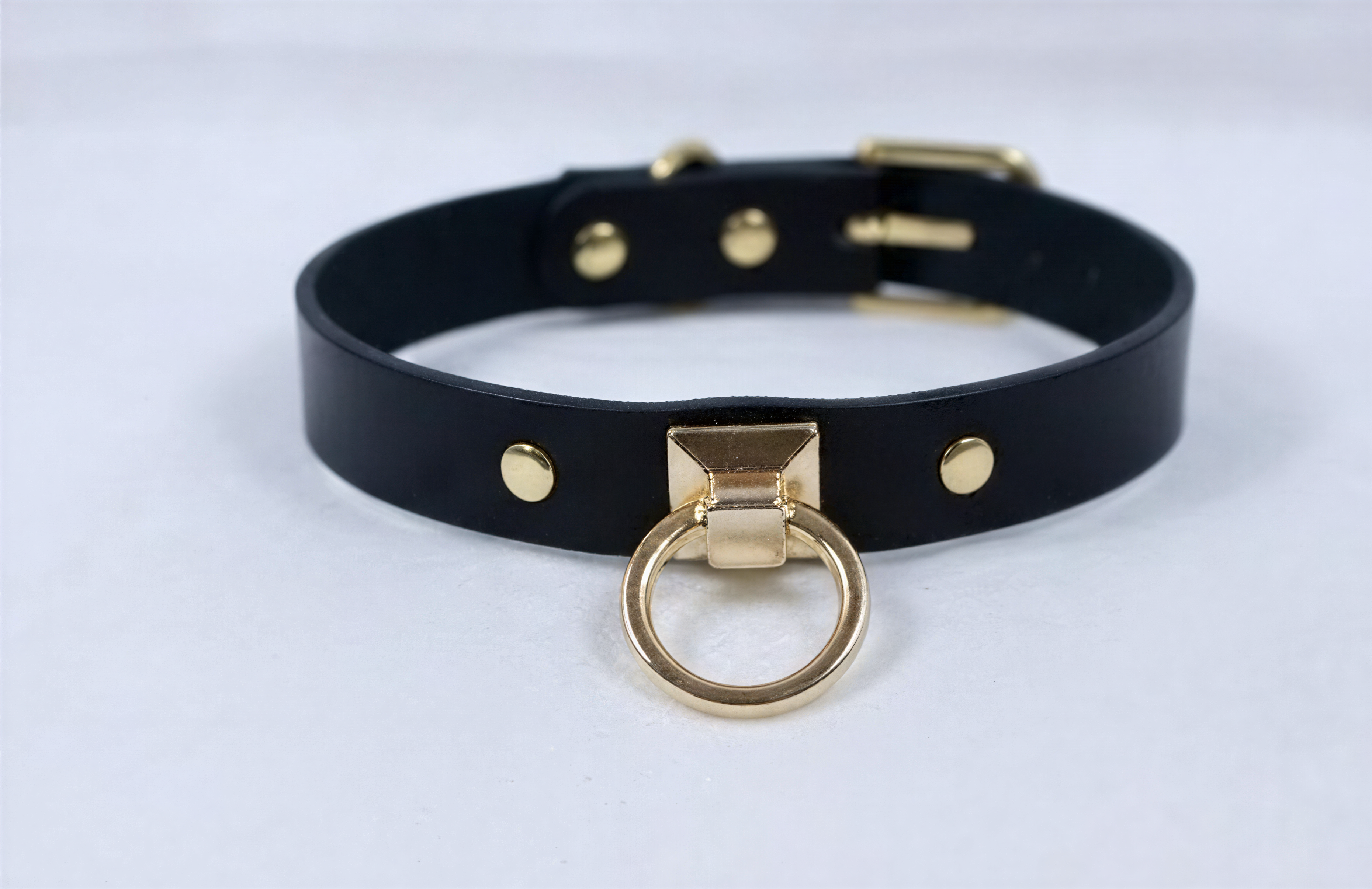 A black leather choker necklace with a gold-colored metal ring and three gold-colored metal studs on the collar.