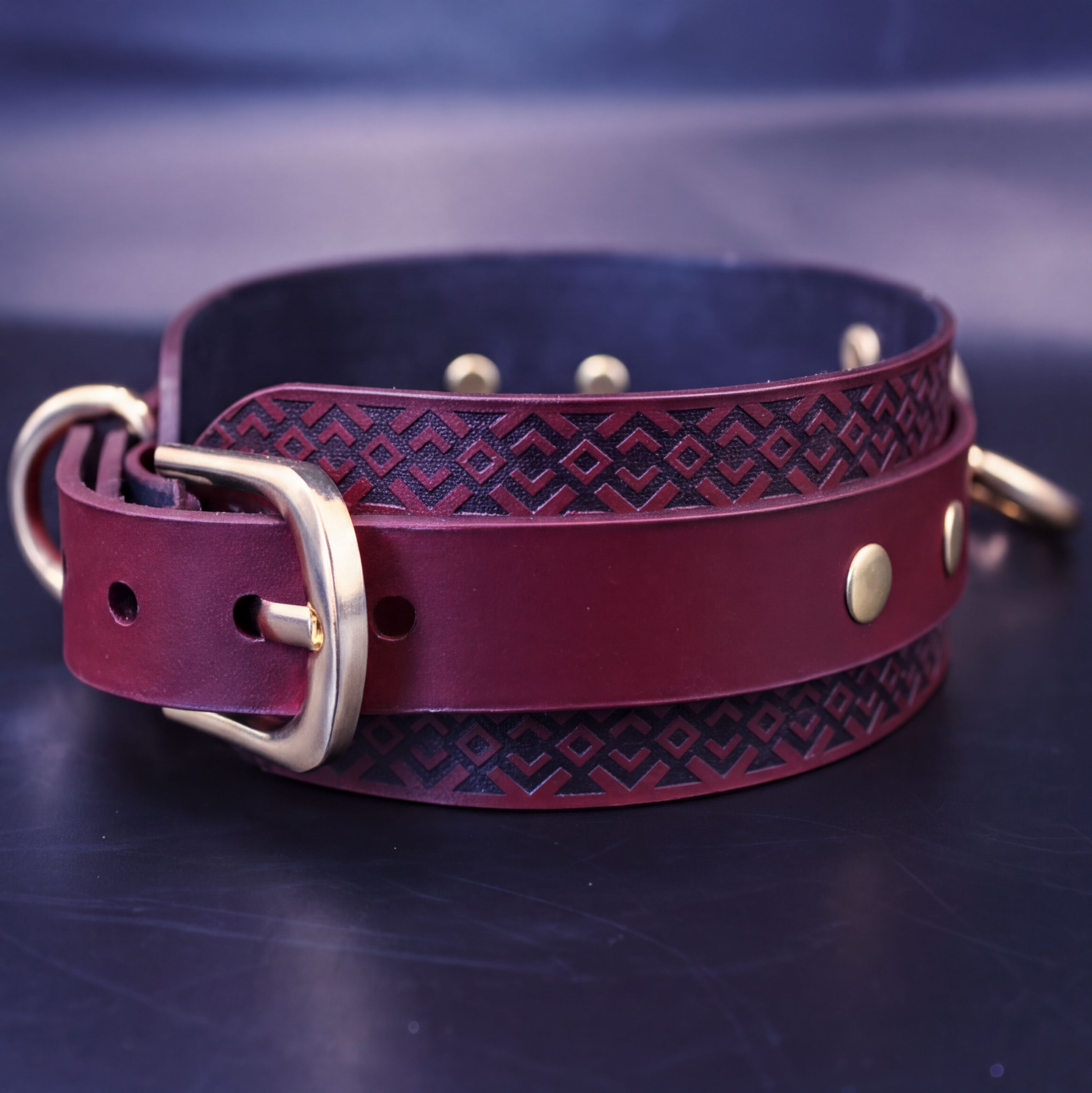 A burgundy leather collar with a geometric pattern and a gold buckle is displayed on a dark surface.