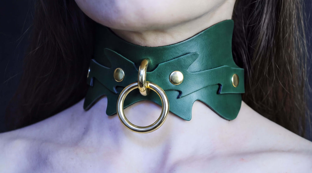 A close-up of a person's neck wearing a green leather choker necklace with a gold ring and two gold studs on the collar.