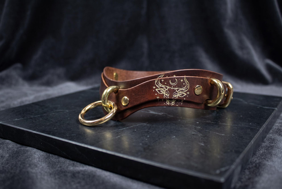 A brown leather strap with gold rings and a skull design on it, resting on a black surface.