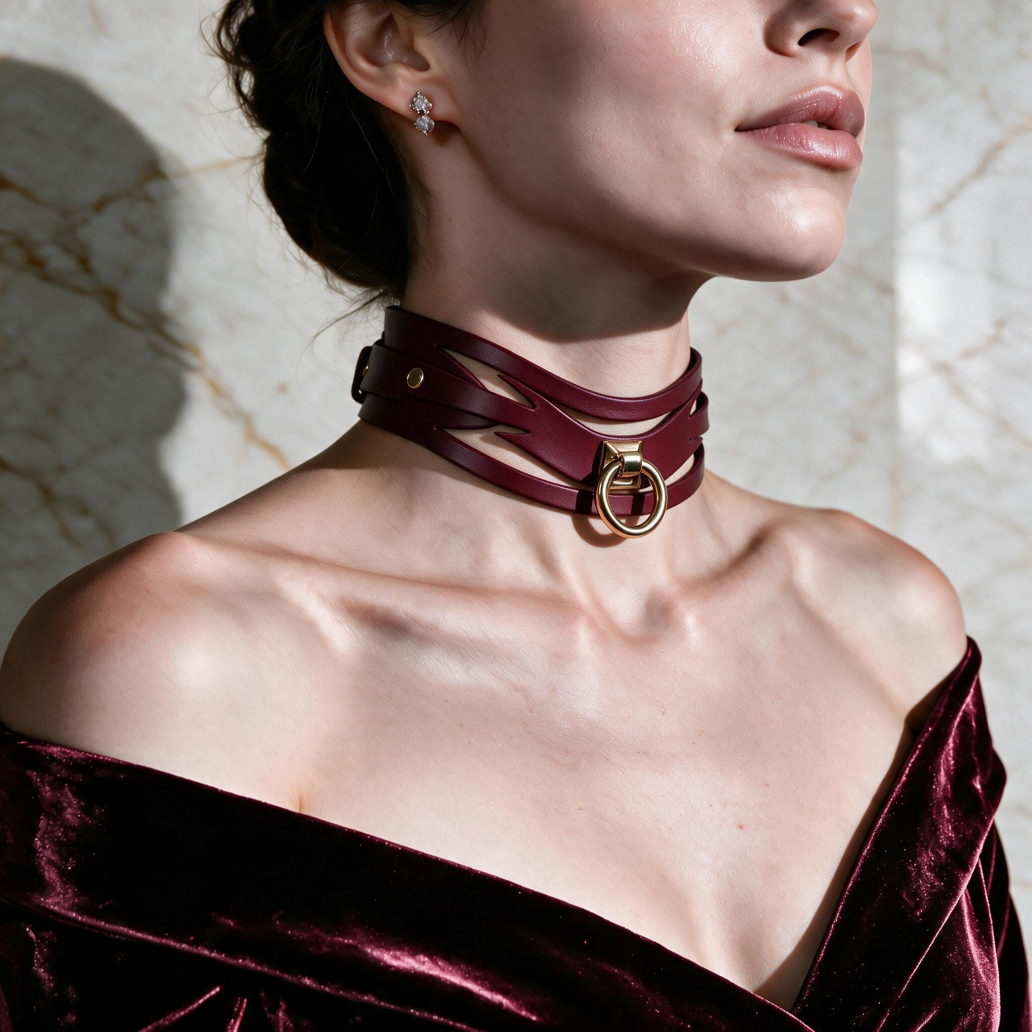 A woman wearing a burgundy choker necklace with a gold buckle, posing against a marble wall.