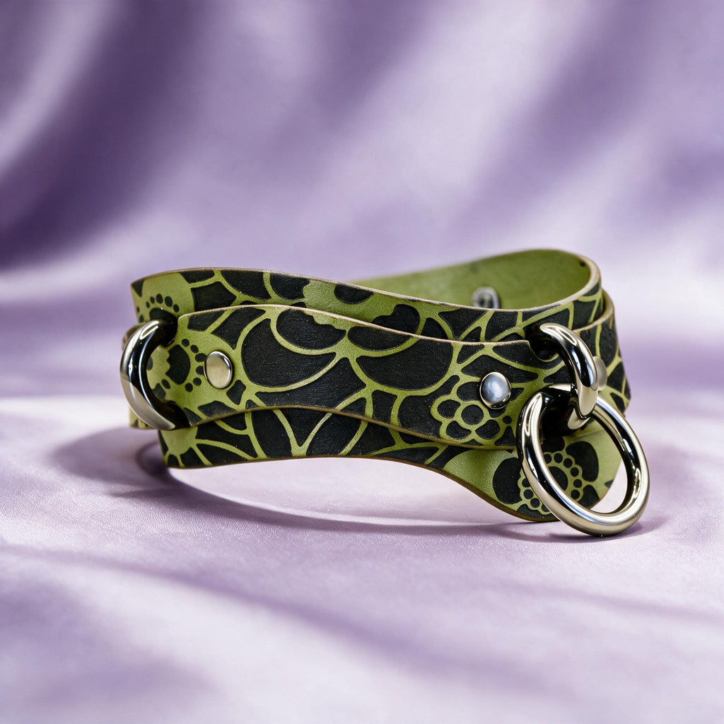 A green and black patterned leather choker necklace with metal rings and a clasp is displayed against a purple background.