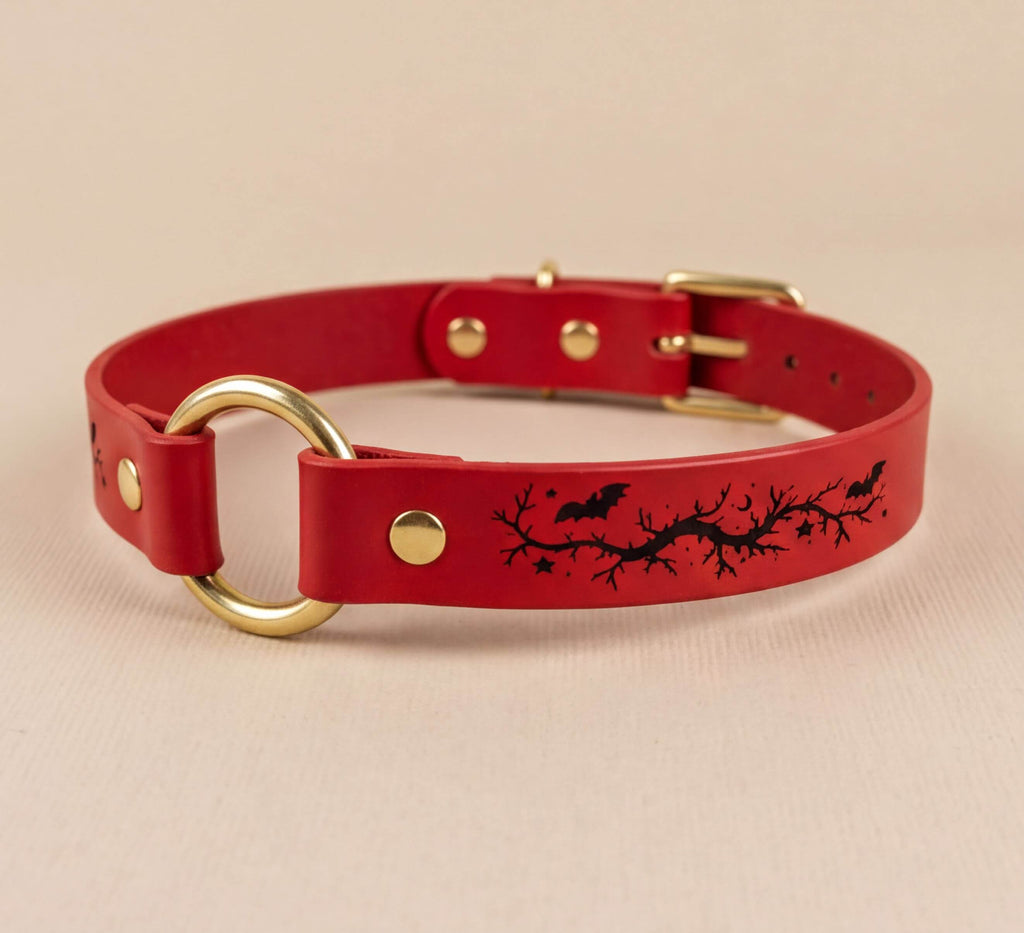 A red leather collar with a gold ring and a black bat and branch design on the front.