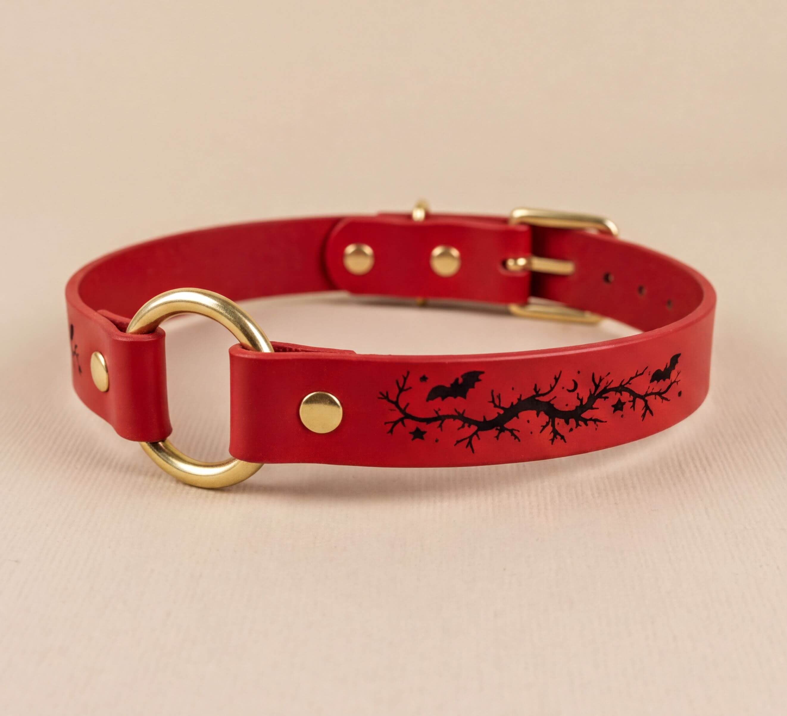 A red leather collar with a gold ring and a black bat and branch design on the front.