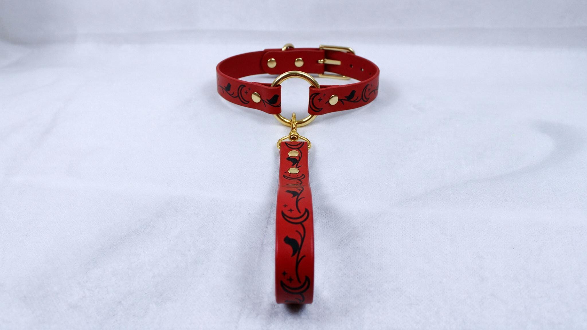 A red leather collar with a gold buckle and a black floral pattern on the strap is displayed against a white background.