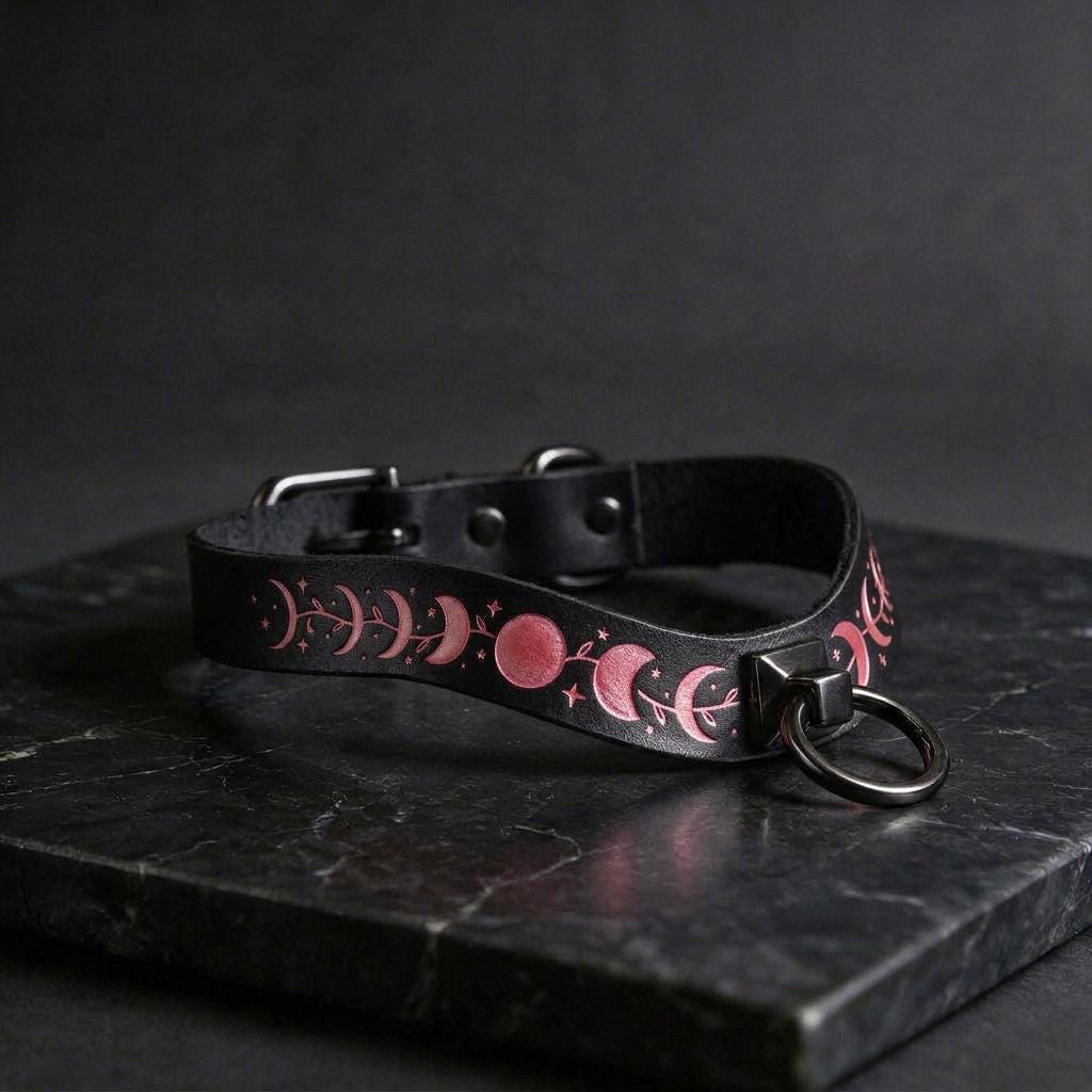 A black leather collar with a pink moon and star design on it, resting on a dark surface.