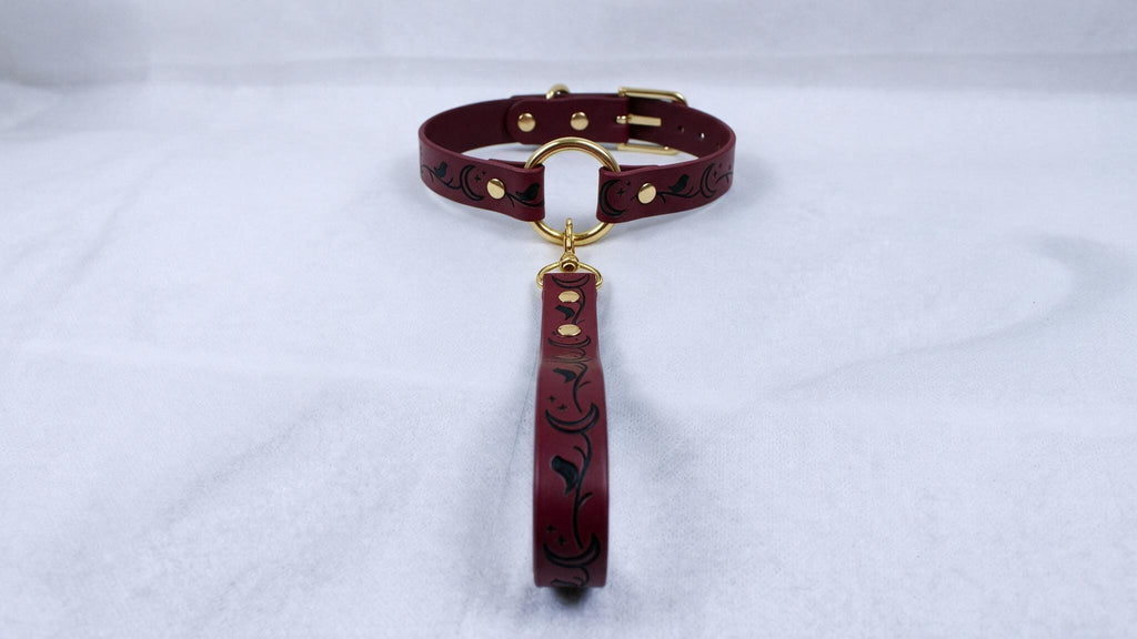 A burgundy leather collar with a gold-colored buckle and a black floral pattern on the strap is displayed on a white surface.