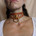 A close-up of a woman's neck and chest area, featuring a choker-style leather necklace with a gold ring.