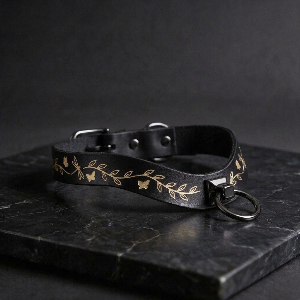 A black leather collar with a leaf and butterfly design, featuring a metal ring and buckle, is displayed on a black marble surface.
