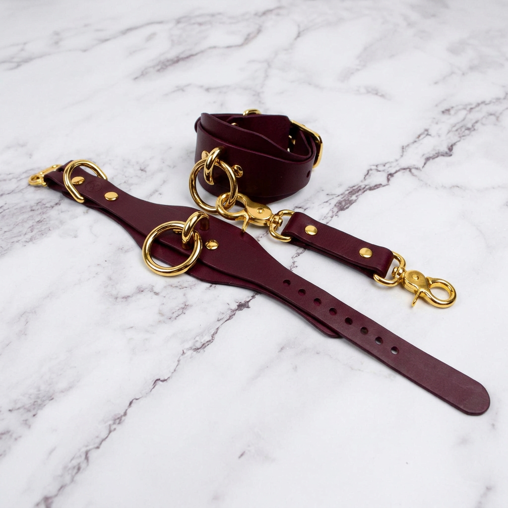 A burgundy leather strap with gold-colored hardware, including rings and buckles, is displayed on a marble surface.