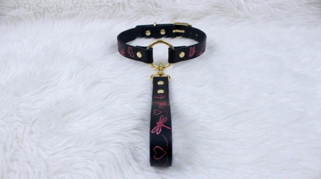 A black leather collar with a gold buckle and a pink and black patterned strap is displayed on a white furry surface.