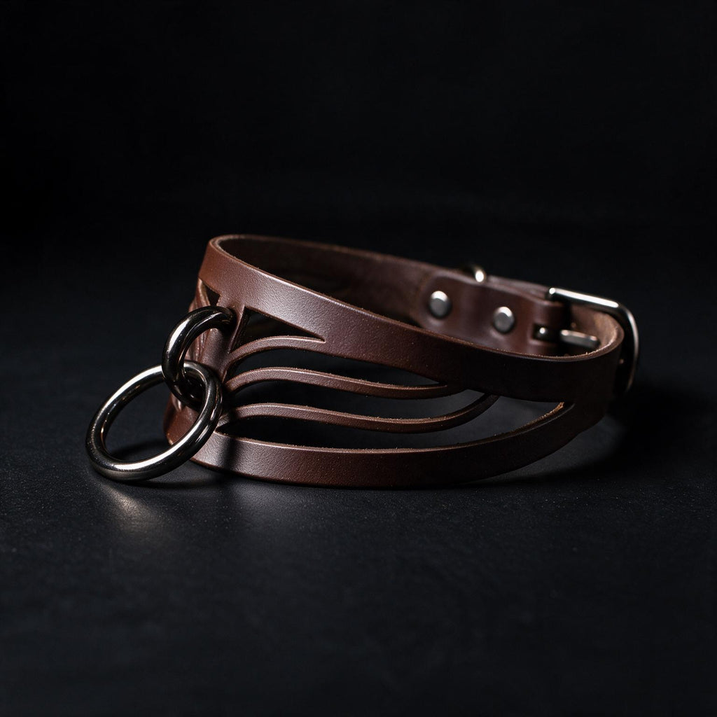 A brown leather choker necklace with metal rings and a buckle is displayed against a dark background.