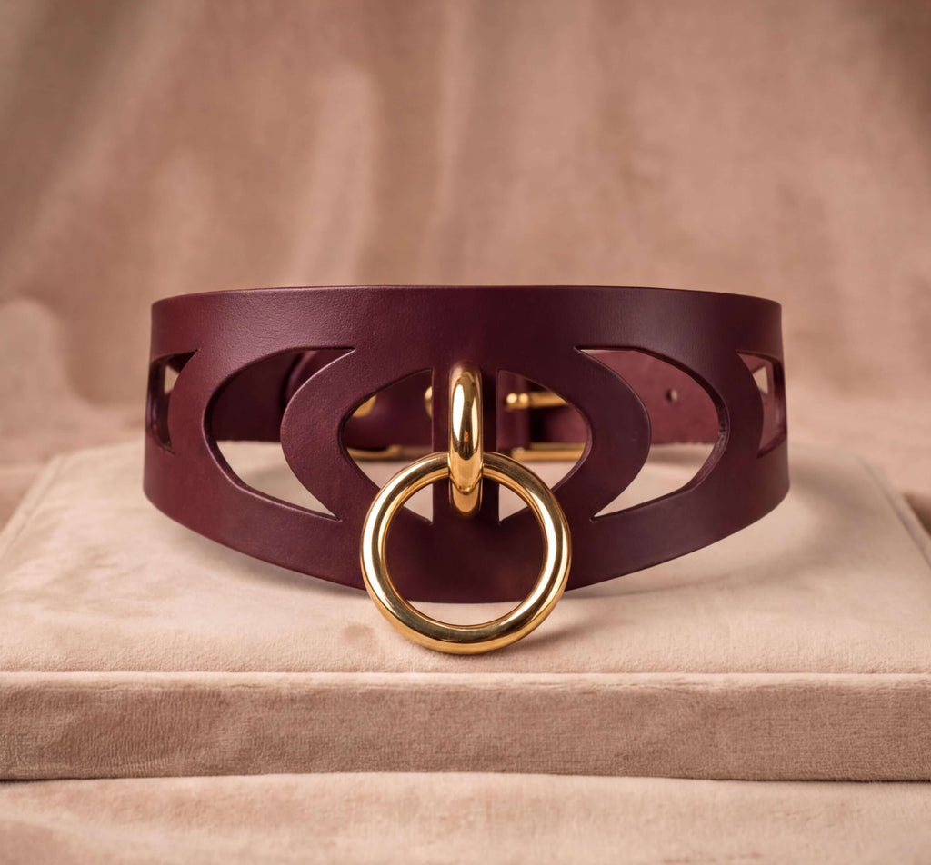 A burgundy leather choker necklace with a gold-colored circular clasp and a cutout design on the front.