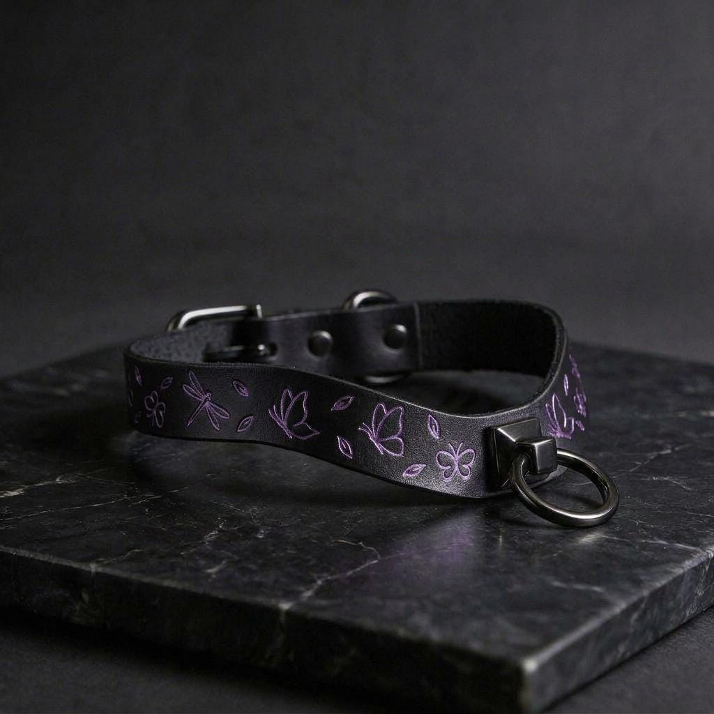 A black leather collar with a silver metal ring and a floral design on it, resting on a dark surface.