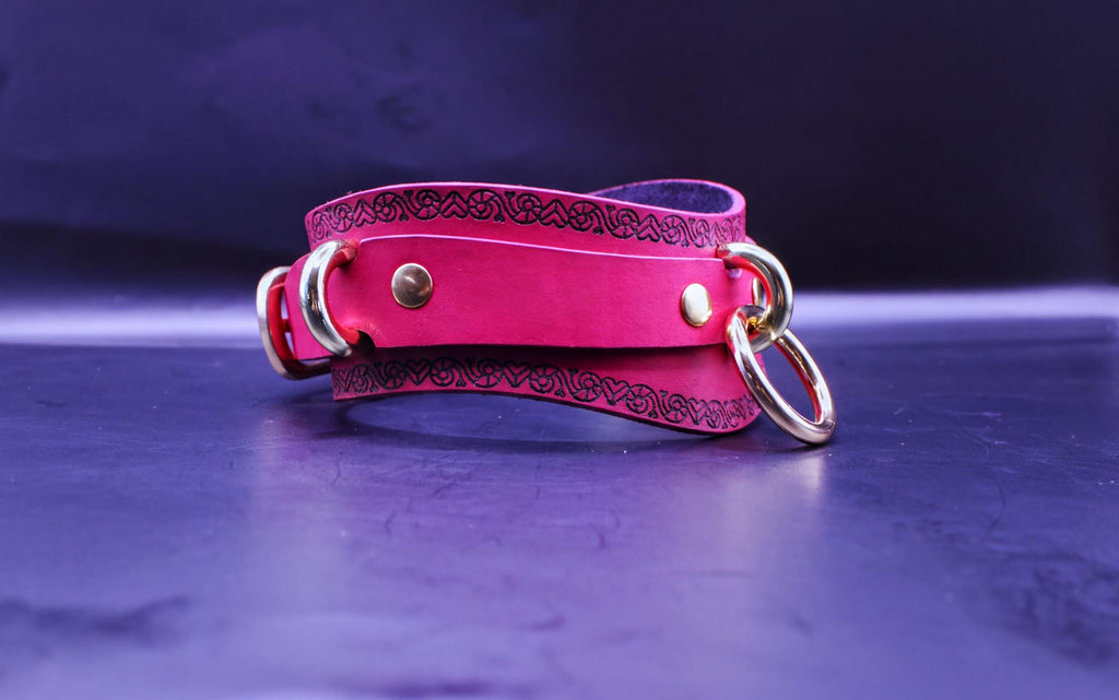 A pink leather choker necklace with a decorative pattern and two metal rings on the front is displayed against a dark purple background.