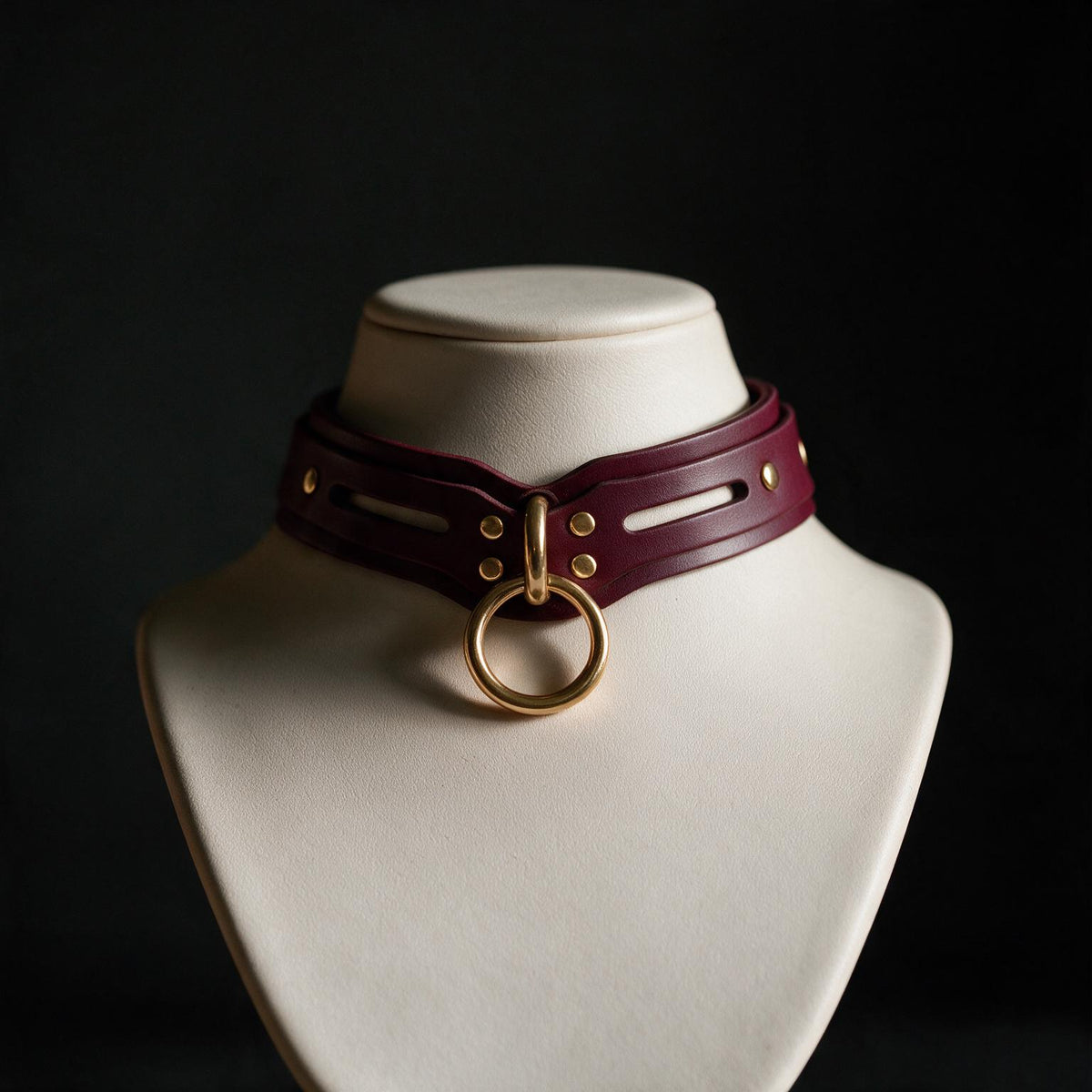 A close-up view of a burgundy leather choker necklace with a gold ring and studs, displayed on a white bust form against a dark background.