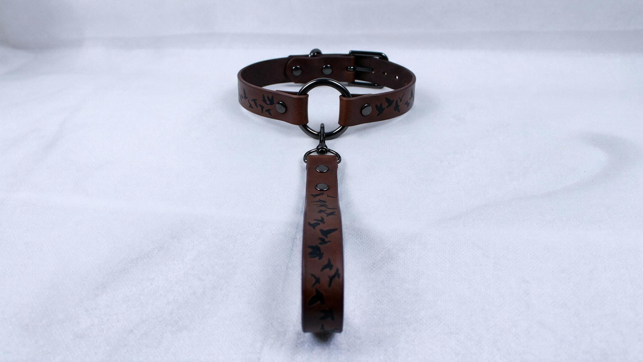 A brown leather collar with a metal buckle and a black handle attached to it, resting on a white surface.