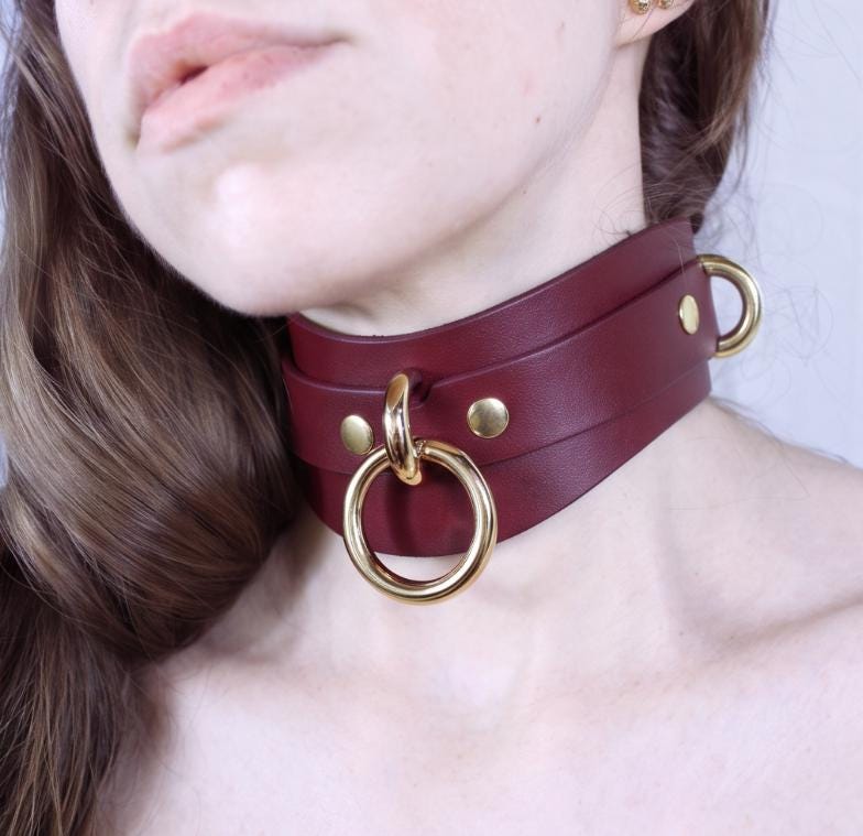 A close-up of a woman's neck wearing a burgundy leather choker necklace with gold-colored metal rings.