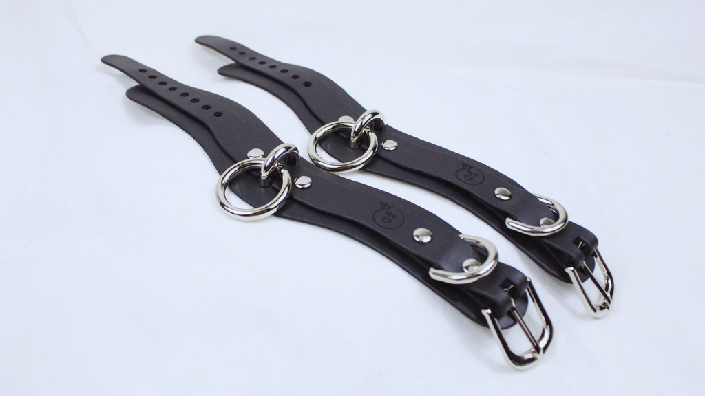 A pair of black leather straps with metal rings and buckles, lying on a white surface.