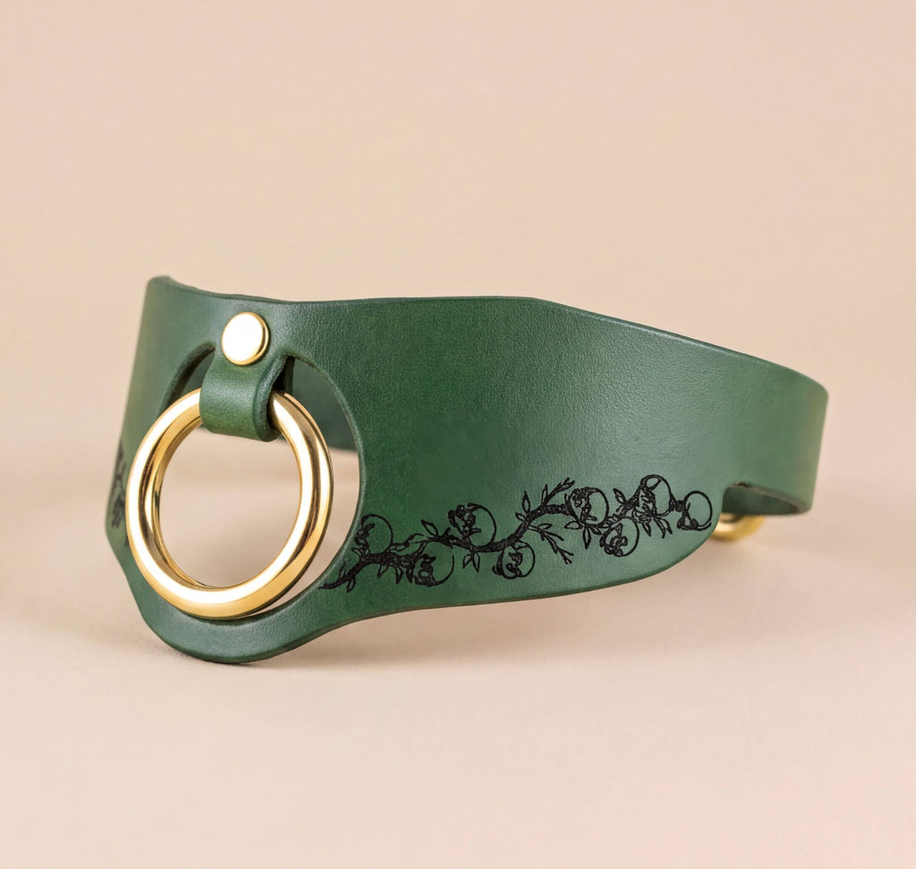 A green leather choker necklace with a gold-colored ring and a black floral design on the front.